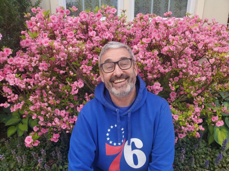 A man with graying hair, a beard, wearing a blue sweatshirt smiling in front of a large pink azalea bush. 